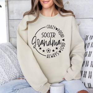 K&ouml;nnte beinhalten: Ein beiger Pullover mit einem schwarzen Kreis-Motiv, auf dem "Crazy Proud Soccer Grandma Always Loud" mit Sternen und einem Fu&szlig;ball steht.