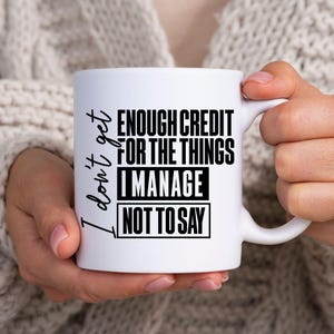May include: White ceramic mug with the text "I don't get enough credit for the things I manage not to say" in black. The mug is held by a person wearing a light-colored sweater.