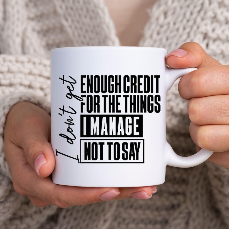 May include: White ceramic mug with the text "I don't get enough credit for the things I manage not to say" in black. The mug is held by a person wearing a light-colored sweater.