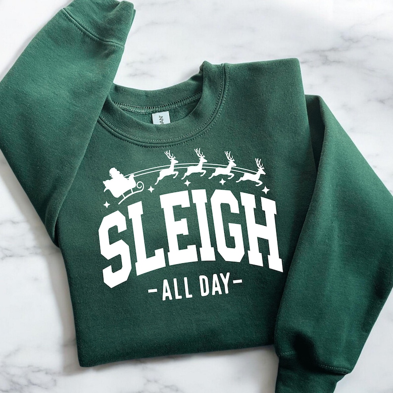 K&ouml;nnte beinhalten: Ein dunkelgr&uuml;ner Pullover mit einem wei&szlig;en Aufdruck eines von Rentieren gezogenen Schlittens mit dem Text "Sleigh - All Day -" auf der Vorderseite.