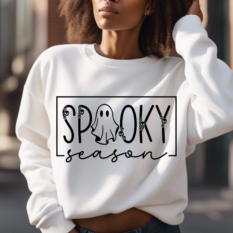 Pu&ograve; includere: Felpa bianca con la scritta nera "Spooky Season". La parola "Spooky" &egrave; in grassetto e "season" &egrave; in corsivo. Piccole grafiche di teschi su entrambi i lati della parola "Spooky".