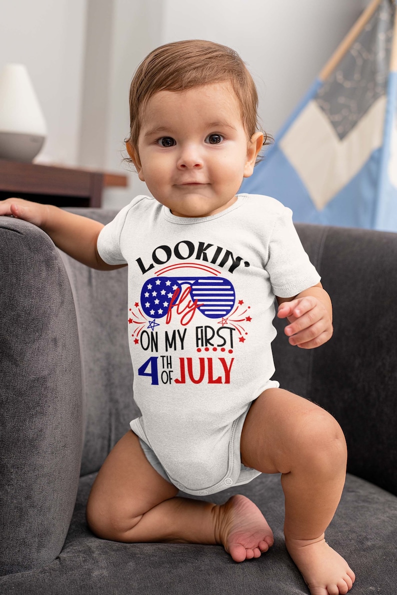 May include: A white baby bodysuit with the text "Lookin' Fly on My First 4th of July" in red, white, and blue. The design includes sunglasses with a stars and stripes pattern. The baby is sitting on a gray couch.
