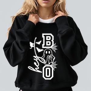 Puede incluir: Sudadera negra con un gr&aacute;fico blanco que dice "Hey Boo". El dise&ntilde;o incluye un fantasma, murci&eacute;lagos y telara&ntilde;as. La letra "B" est&aacute; en un estilo de bloque, y la "O" es un cero.