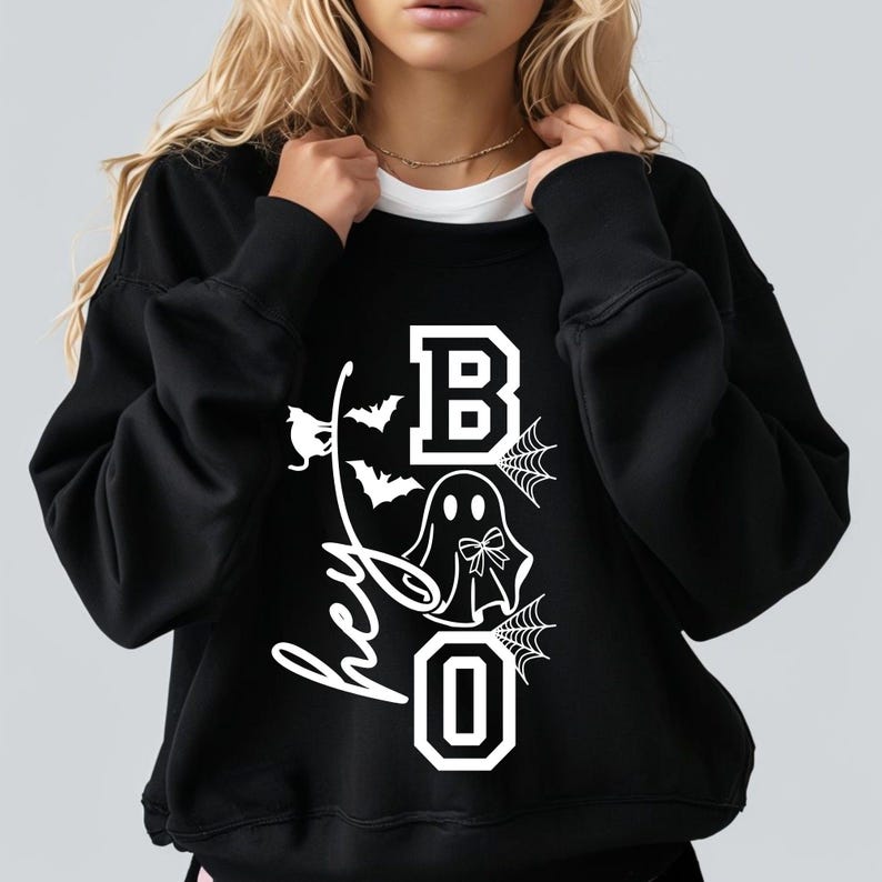 Puede incluir: Sudadera negra con un gr&aacute;fico blanco que dice "Hey Boo". El dise&ntilde;o incluye un fantasma, murci&eacute;lagos y telara&ntilde;as. La letra "B" est&aacute; en un estilo de bloque, y la "O" es un cero.