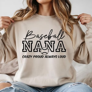 May include: Beige sweatshirt with the text "Baseball NANA" in black script and block letters, featuring a baseball graphic. The phrase "CRAZY PROUD ALWAYS LOUD" is printed below the baseball graphic.