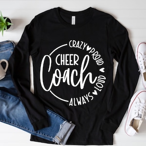 May include: Black long-sleeved shirt with the words "Cheer Coach" in a white circular design. The words "Crazy, Proud, Loud, Always" are also in white. The shirt is on a white surface with blue jeans, a mug, and white trainers.