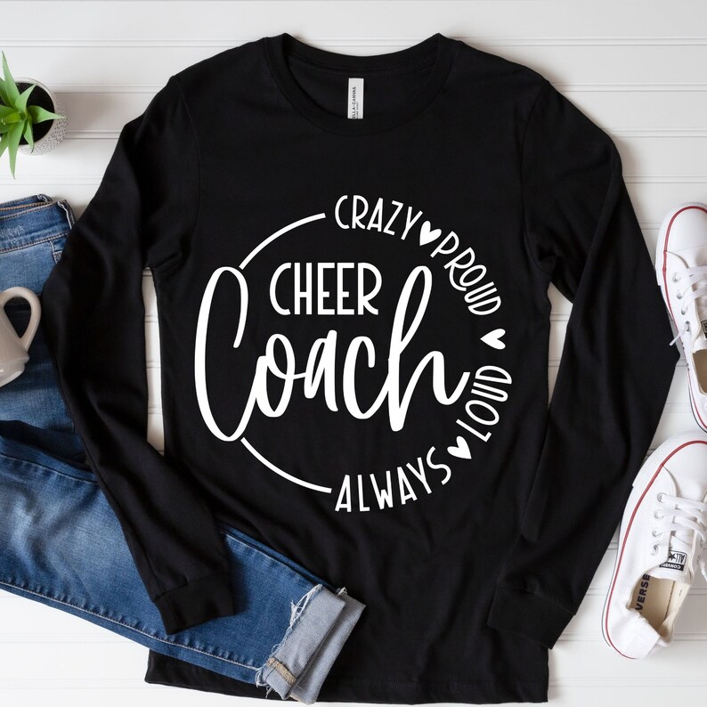 May include: Black long-sleeved shirt with the words "Cheer Coach" in a white circular design. The words "Crazy, Proud, Loud, Always" are also in white. The shirt is on a white surface with blue jeans, a mug, and white trainers.