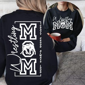Puede incluir: Sudadera negra con las palabras "Wrestling MOM" en letras blancas y en escritura. La sudadera tambi&eacute;n presenta un gr&aacute;fico de dos luchadores y las palabras "Crazy, Proud, Always, Loud."