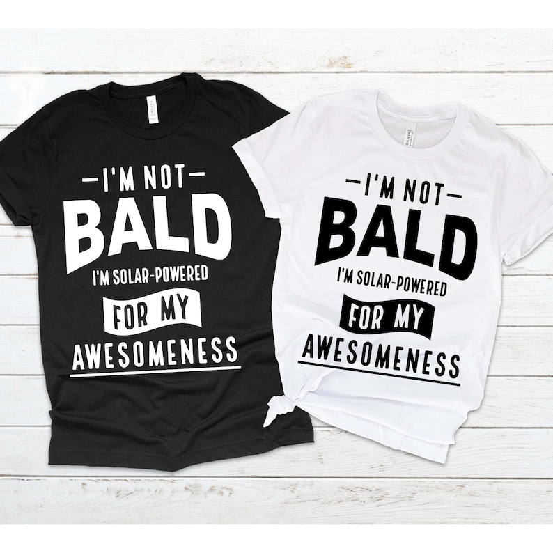 May include: Two t-shirts with a black and white graphic design. The text on the shirts reads "I'm not bald, I'm solar-powered for my awesomeness".