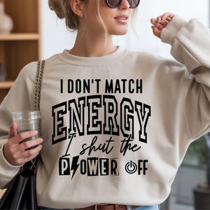 K&ouml;nnte beinhalten: Beiges Sweatshirt mit dem schwarzen Text "I DON'T MATCH ENERGY I shut the POWER OFF". Das Sweatshirt hat einen Rundhalsausschnitt und lange &Auml;rmel. Das Bild zeigt eine Person, die das Sweatshirt tr&auml;gt.