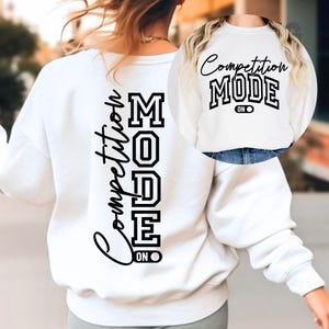 May include: A white sweatshirt featuring the text "Competition Mode" in black. The words are arranged vertically on the back and horizontally on the front. The sweatshirt has a crew neck design.