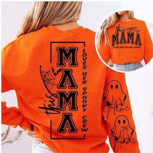 Pode incluir: Camisola laranja com texto e gráficos pretos. As costas apresentam a palavra "MAMA" em letras grandes, com "This" e "Loves Her Spooky Crew" em letras menores. As mangas têm gráficos de fantasmas.