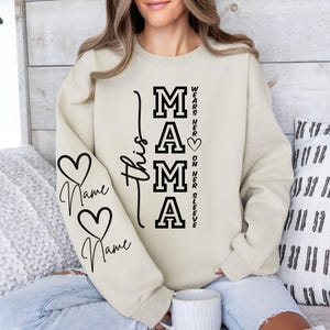 Op de afbeelding: Crèmekleurige sweatshirt met het woord "MAMA" in vette zwarte letters. De mouw heeft twee harten met het woord "Name" eronder. De tekst "This" en "Wears her heart on her sleeve" staan ook op de sweatshirt.