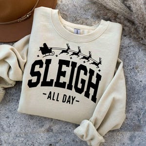 K&ouml;nnte beinhalten: Ein beiger Pullover mit einem schwarzen Aufdruck von Santa Claus in einem Schlitten, der von Rentieren gezogen wird. Der Text "SLEIGH -ALL DAY- " ist unter dem Aufdruck gedruckt.