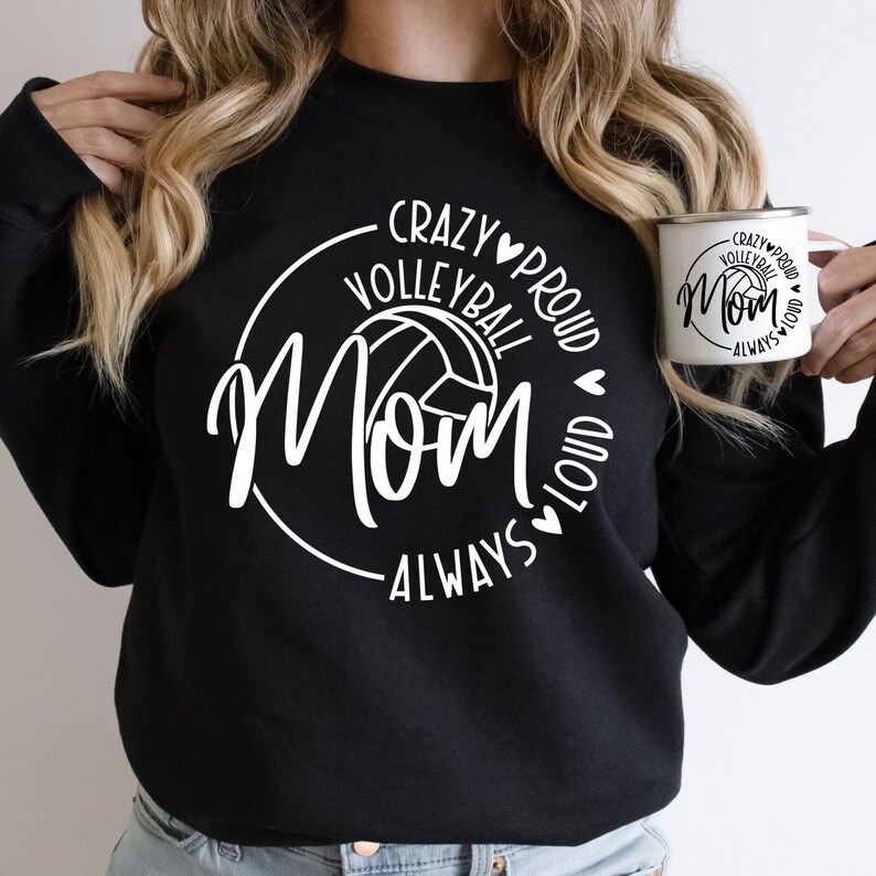 May include: Black sweatshirt with white text that reads "Crazy Proud Volleyball Mom Always Loud" with a volleyball graphic in the center. The sweatshirt is worn by a person with long blonde hair. A white mug with the same text and graphic is in the person's hand.