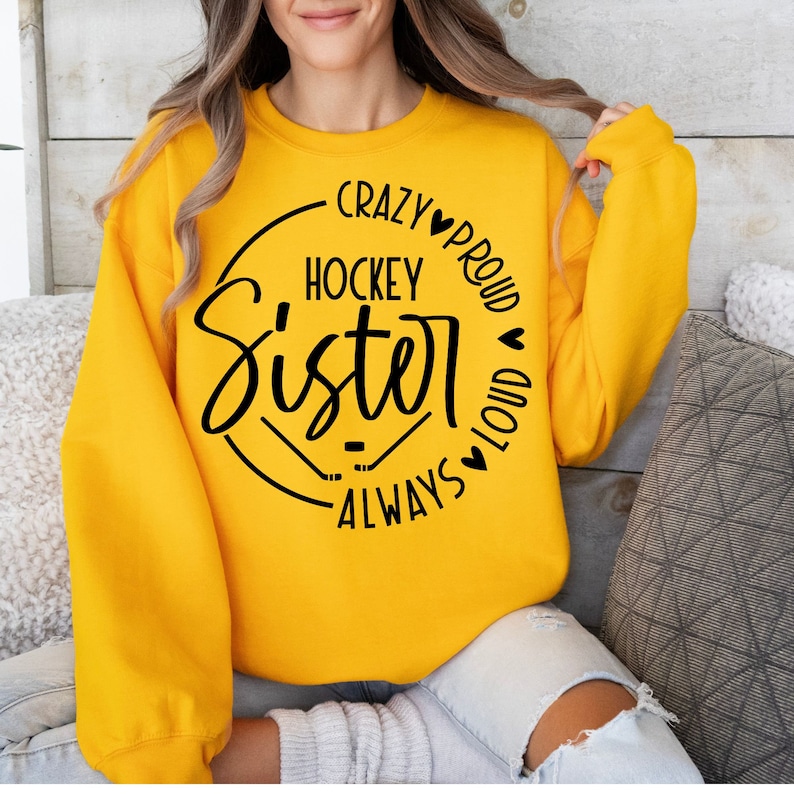 Puede incluir: Sudadera amarilla con un gr&aacute;fico circular negro que dice "Crazy Hockey Sister Always Proud and Loud" con corazones y palos de hockey.