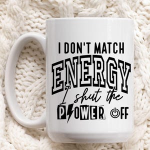 K&ouml;nnte beinhalten: Wei&szlig;e Keramiktasse mit schwarzem Aufdruck "I DON'T MATCH ENERGY I shut the POWER OFF". Die Tasse hat einen gro&szlig;en Henkel und steht vor einem wei&szlig;en, strukturierten Hintergrund.