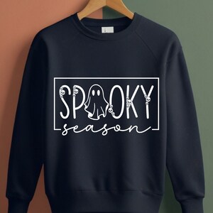 Pu&ograve; includere: Felpa blu navy con le parole bianche "SP00KY season" all'interno di un rettangolo bianco. La parola "SP00KY" presenta grafiche di fantasmi e teschi. La felpa &egrave; appesa a una gruccia di legno.