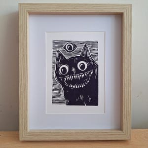 May include: Framed black and white art print of a grinning cat with large eyes and sharp teeth, and a floating eye above. The artwork is in a light wood frame with a white mount.