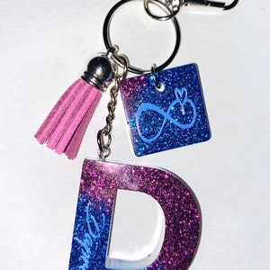 Peut inclure: Un porte-clés composé d'une grande lettre D et d'une breloque carrée, tous deux avec une finition à paillettes bleues et violettes. La lettre D porte l'inscription Daphne. Un pompon en daim rose et un mousqueton argenté sont fixés à l'anneau.