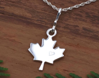 May include: A silver-colored pendant necklace featuring a maple leaf design with a small heart detail. The pendant hangs from a delicate silver chain. The necklace is displayed on a wooden surface with green foliage.
