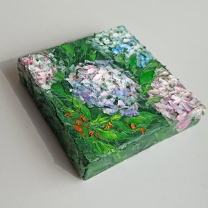 May include: A small painting of a cluster of hydrangeas in shades of pink, white, and blue, with green leaves and red berries. The painting is on a canvas and has a textured surface.