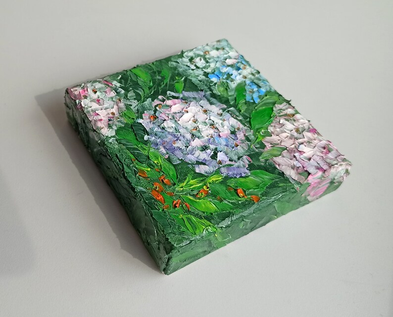 May include: A small painting of a cluster of hydrangeas in shades of pink, white, and blue, with green leaves and red berries. The painting is on a canvas and has a textured surface.