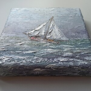 May include: A small painting on canvas depicting a sailboat with white sails on a choppy sea. The sky is a light gray with hints of blue. The painting is done in a textured style, giving the impression of brushstrokes.