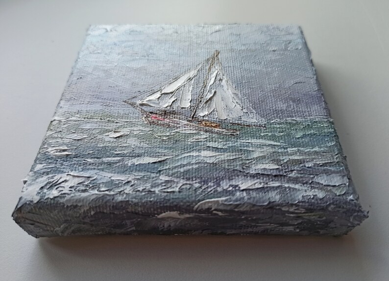 May include: A small painting on canvas depicting a sailboat with white sails on a choppy sea. The sky is a light gray with hints of blue. The painting is done in a textured style, giving the impression of brushstrokes.