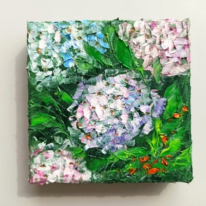 May include: A close-up of a small oil painting depicting a cluster of hydrangeas in shades of pink, blue, and white. The flowers are surrounded by lush green leaves and a few red accents.