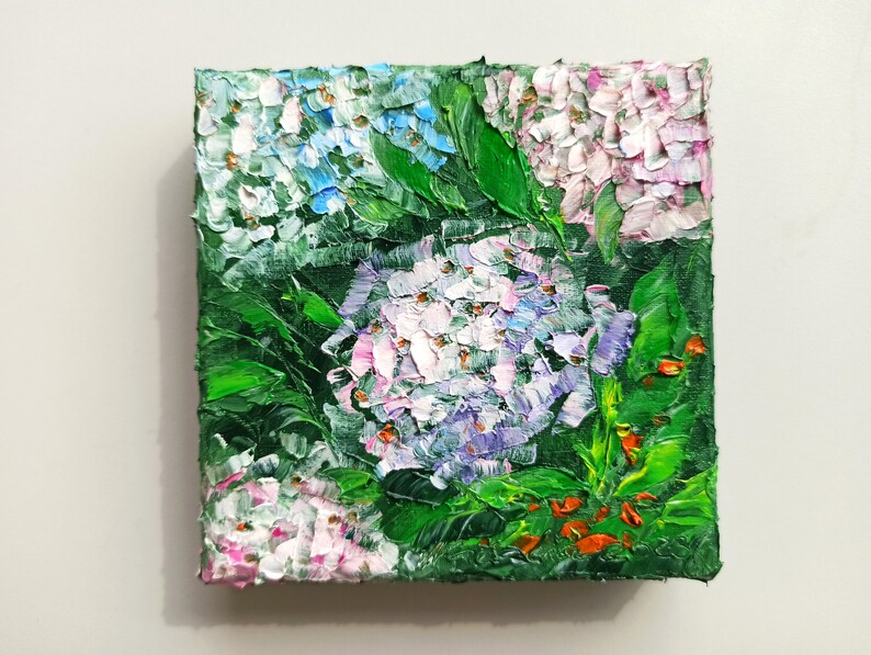 May include: A close-up of a small oil painting depicting a cluster of hydrangeas in shades of pink, blue, and white. The flowers are surrounded by lush green leaves and a few red accents.