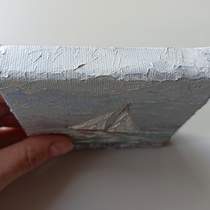 May include: A small, white canvas painting of a sailboat on the water. The painting is done in a loose, impressionistic style, with visible brushstrokes. The sailboat is white with a red hull, and the water is a light blue.