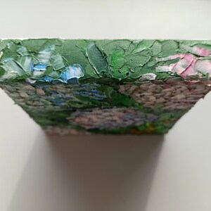 May include: A small, square canvas painting depicting a cluster of pink, blue, and white flowers with green leaves. The painting is done in a textured style, with visible brushstrokes.