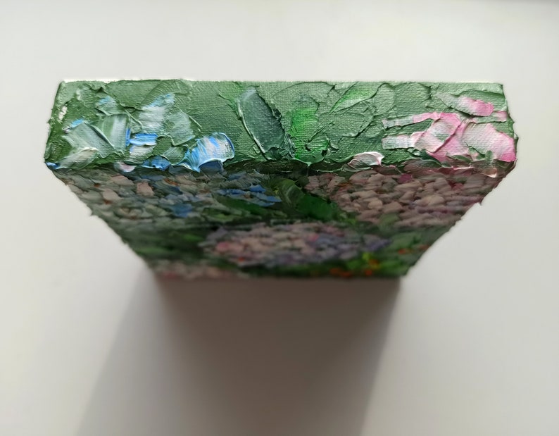 May include: A small, square canvas painting depicting a cluster of pink, blue, and white flowers with green leaves. The painting is done in a textured style, with visible brushstrokes.