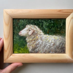 May include: An oil painting of a white sheep with textured brushstrokes, set against a dark green foliage background. The artwork is enclosed in a light-colored, natural wood frame with a visible grain, featuring the year 2025 signed in the corner.