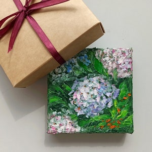 May include: A small painting of a cluster of pink and blue hydrangeas with green leaves. The painting is on a canvas and is framed in a brown wooden frame.