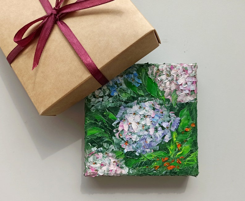 May include: A small painting of a cluster of pink and blue hydrangeas with green leaves. The painting is on a canvas and is framed in a brown wooden frame.