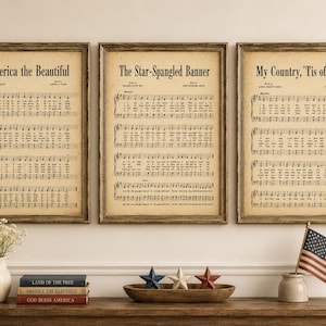 Pode incluir: Três impressões emolduradas de partituras patrióticas: "America the Beautiful", "The Star-Spangled Banner" e "My Country, 'Tis of Thee". As impressões são exibidas acima de uma mesa de madeira com itens decorativos, incluindo livros e uma pequena bandeira americana.