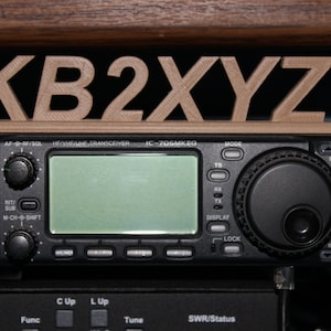 May include: A black Icom IC-706MKEG HF/VHF/UHF transceiver with a wooden nameplate that reads "KB2XYZ" on top. The transceiver has a digital display, a knob for tuning, and several buttons for different functions.