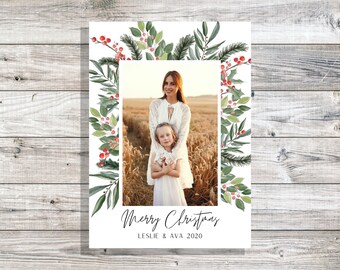 Peut inclure: Une carte de Noël avec une illustration aquarelle de feuilles vertes et de baies rouges entourant une photo d'une mère et de sa fille. Le texte "Merry Christmas" et "Leslie & Ava 2020" est imprimé sous la photo.