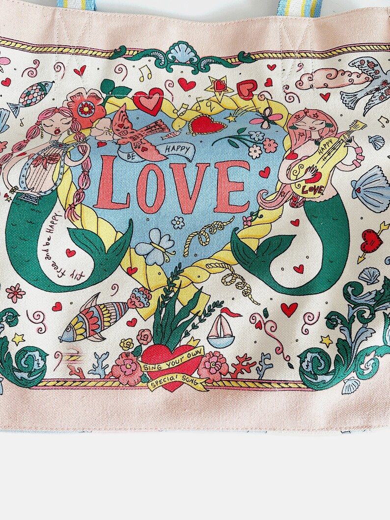 May include: A canvas tote bag with a whimsical design featuring mermaids, hearts, and the word "LOVE" in large letters. The bag has a light pink border and handles with blue and yellow stripes. The overall theme is romantic and nautical.