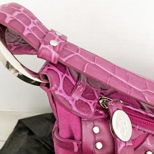 May include: Close-up of a magenta handbag with a crocodile-skin texture. The bag features a silver handle and zipper, with silver rivets and a silver oval tag with the initials "DP". The bag is made of a combination of materials.