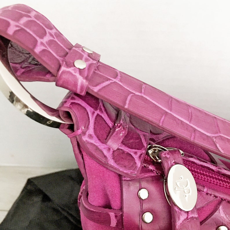 May include: Close-up of a magenta handbag with a crocodile-skin texture. The bag features a silver handle and zipper, with silver rivets and a silver oval tag with the initials "DP". The bag is made of a combination of materials.