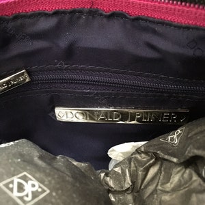 May include: Interior view of a dark pink handbag with a navy blue lining. The bag features a silver zipper and a silver metal plate with the brand name "DONALD J PLINER". The bag is partially obscured by black tissue paper.