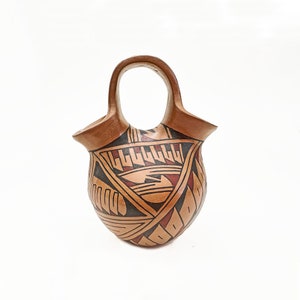 May include: Handmade pottery vessel with a handle. The earthenware pot features a light brown base with geometric patterns in black and red. The handle is a continuation of the pot's rim, creating a unique, functional design.