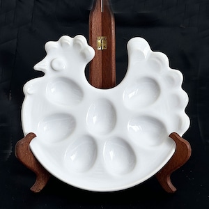 May include: A white ceramic egg serving platter in the shape of a hen, with six egg-shaped indentations. The platter is displayed on a wooden stand. The hen-shaped platter is a decorative and functional kitchen item.