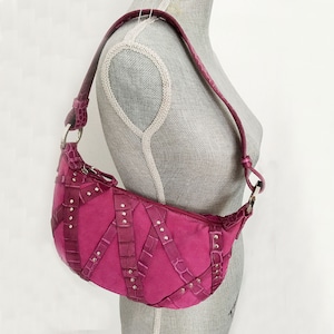 May include: A fuchsia-colored shoulder bag with a curved shape. The bag features a suede-like body with crocodile-textured leather straps and silver-colored stud accents. The strap is also made of crocodile-textured leather.
