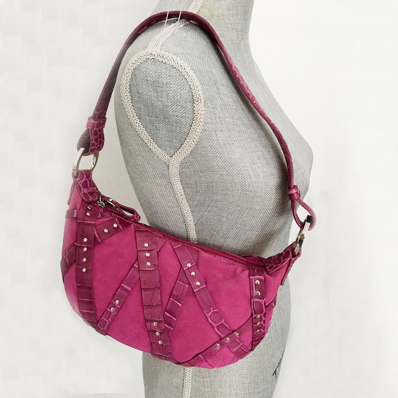 May include: A fuchsia-colored shoulder bag with a curved shape. The bag features a suede-like body with crocodile-textured leather straps and silver-colored stud accents. The strap is also made of crocodile-textured leather.