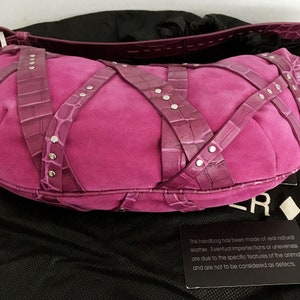 May include: A fuchsia suede handbag with crocodile-textured straps crisscrossing the body. Silver-tone rivets secure the straps. The bag has a rounded shape and a matching strap. The bag is on a black surface.