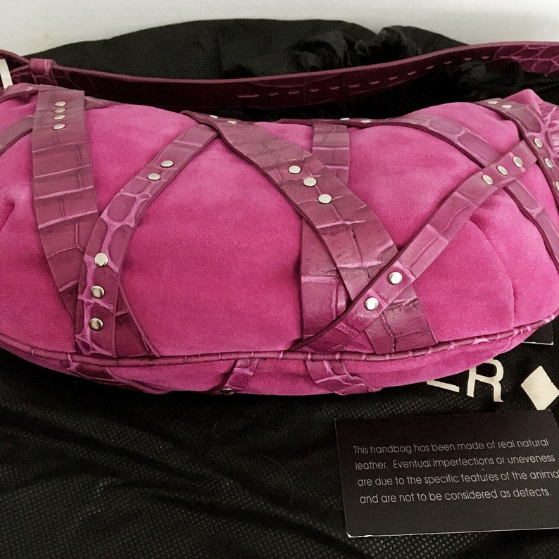 May include: A fuchsia suede handbag with crocodile-textured straps crisscrossing the body. Silver-tone rivets secure the straps. The bag has a rounded shape and a matching strap. The bag is on a black surface.